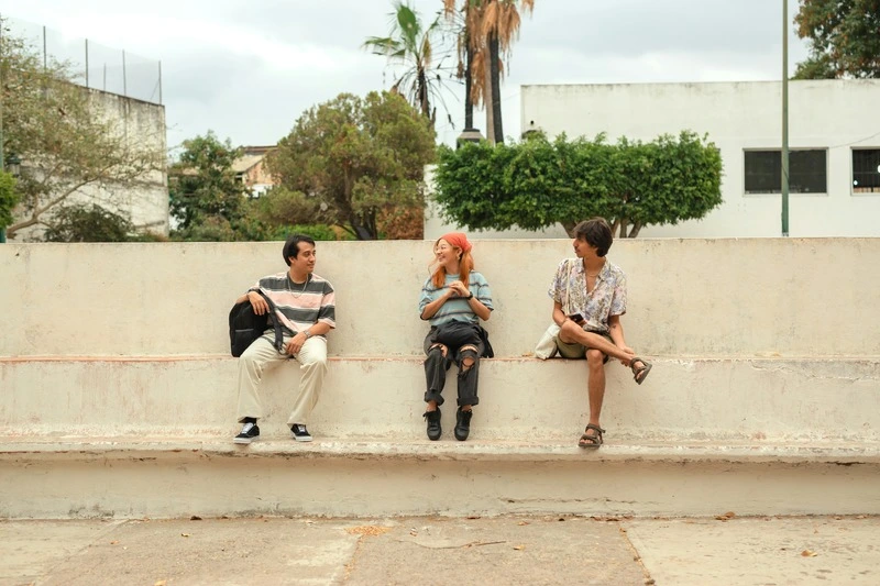 imagem mostra tres pessoas sentada em um banco de concreto distantes entre si com arvores no fundo.