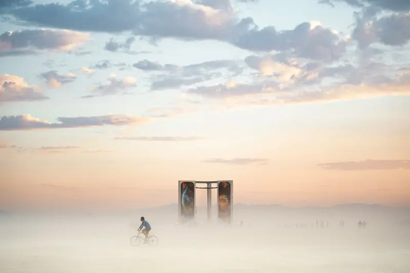 Profissionais de marketing como deuses. Pessoa andando de bicicleta em paisagem com névoa no chão, pessoa s ao fundo e céu com poucas nuvens. Destaque para mobiliário urbano. 